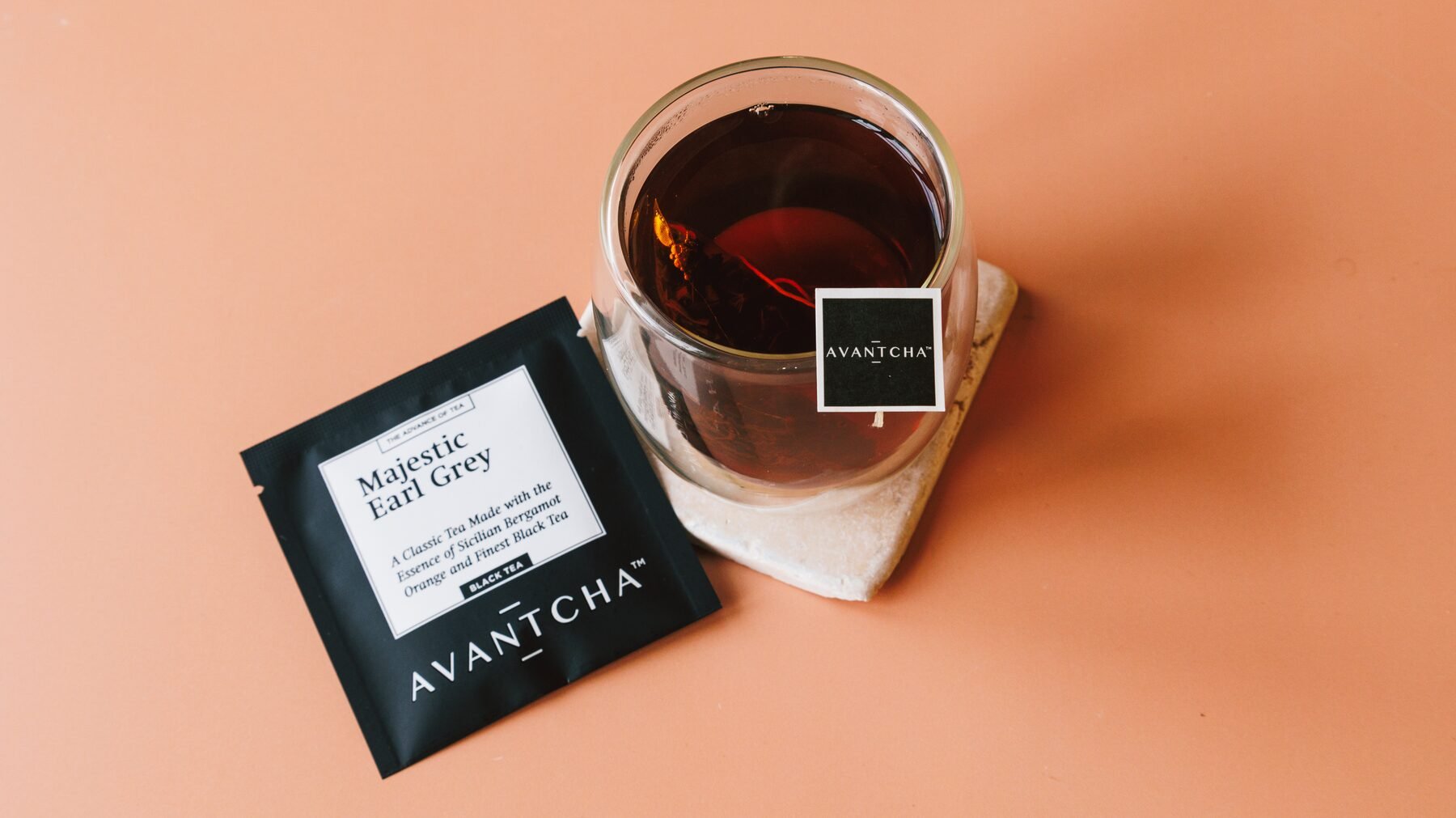 Majestic Earl Grey sachet beside a double-walled glass of brewed tea on a soft terracotta surface